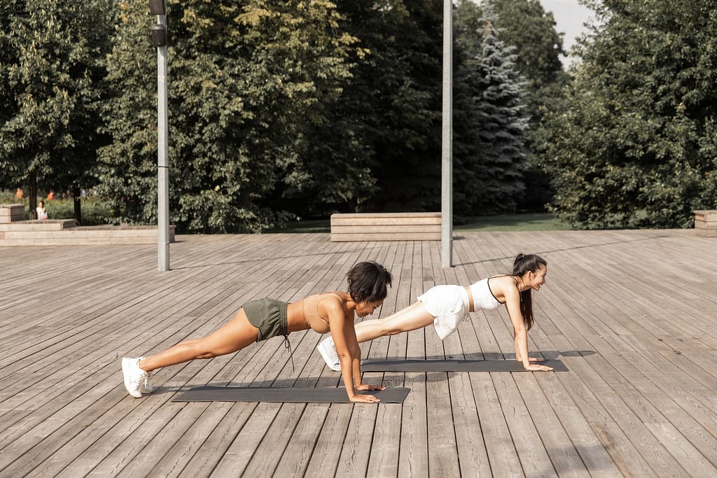 Two women practicing a plank exercise on a wooden deck outdoors.